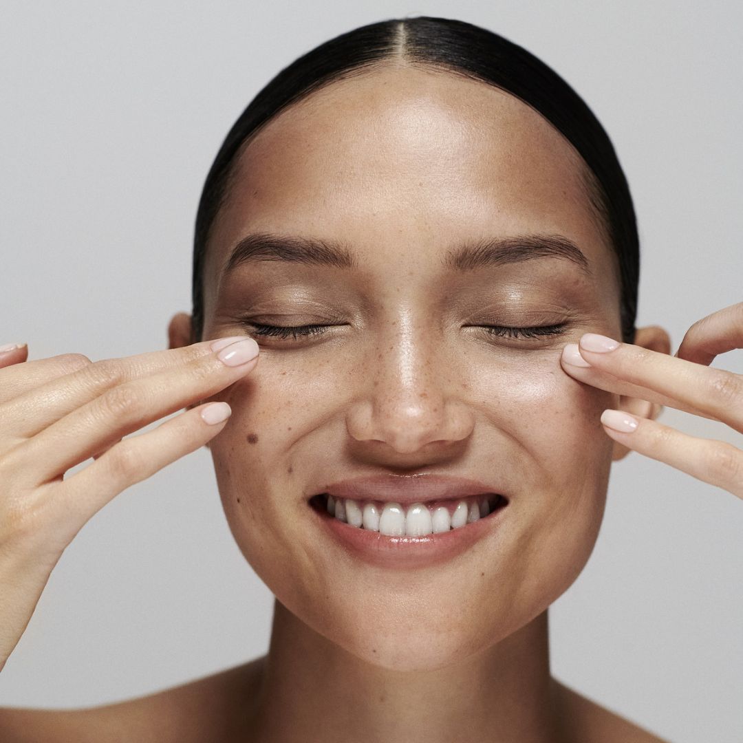 Model smiling with her eyes closed, with both hands' fingertips on her undereye area